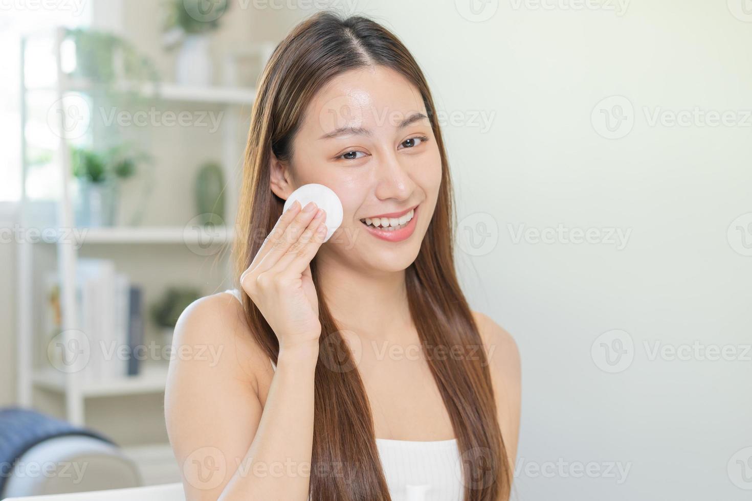 Happy beauty, beautiful asian young woman, girl smiling, looking in mirror, holding cotton pad, applying lotion by wipe on her face, removing makeup before shower at home. Skin care routine people. photo