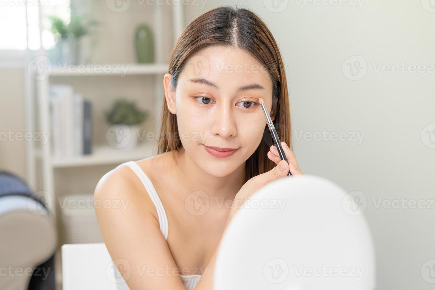 Happy beauty concept, cute asian young woman, girl smile, make up face by an eyeshadow brush on her eyes, looking into mirror in morning routine at home. People look with natural fashion style. photo