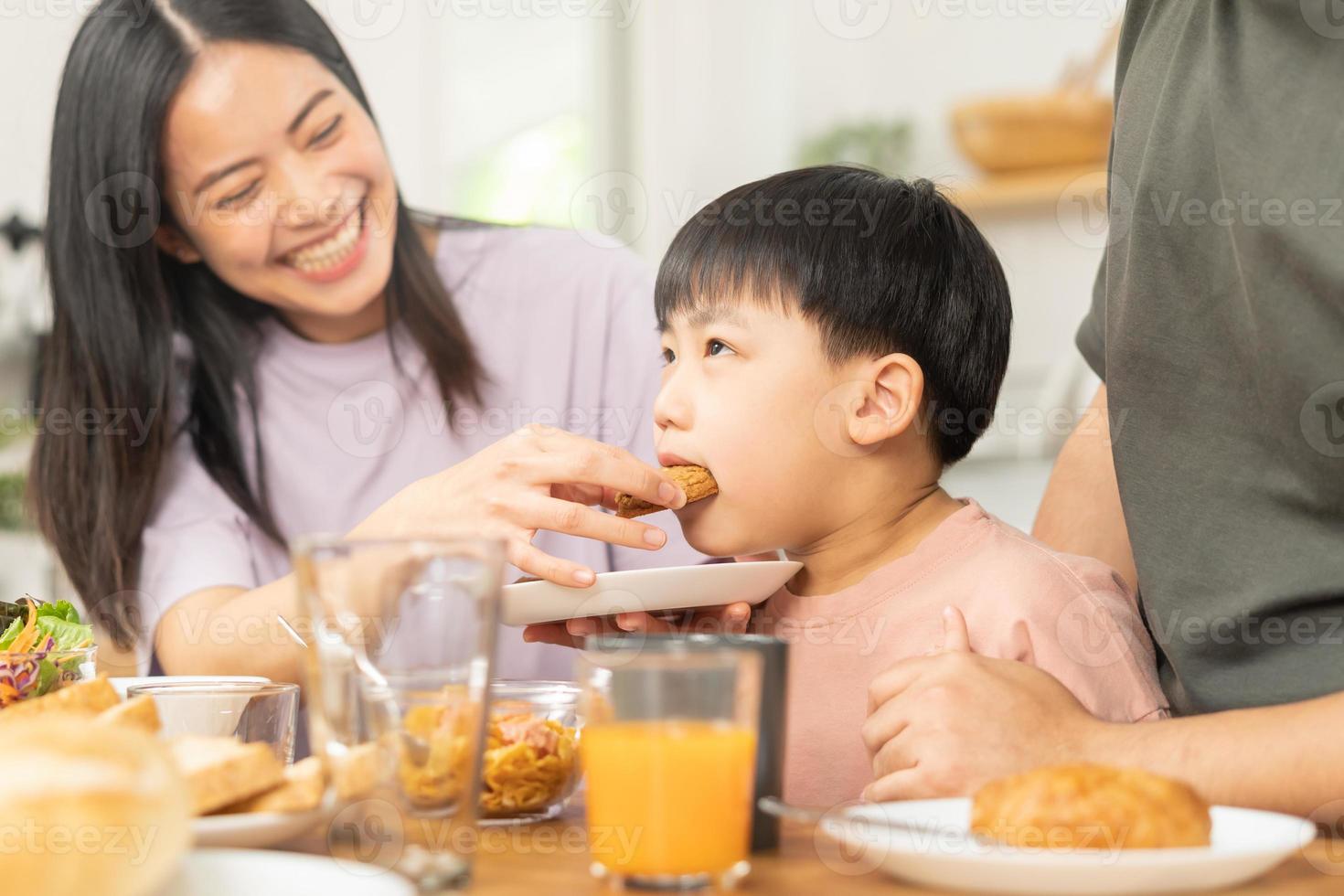 contento refresco familia desayuno en mañana, asiático joven padre padre, madre y pequeño linda chico, niño teniendo comida en cocina comiendo juntos a hogar. alegre, disfrutar Cocinando gente. foto