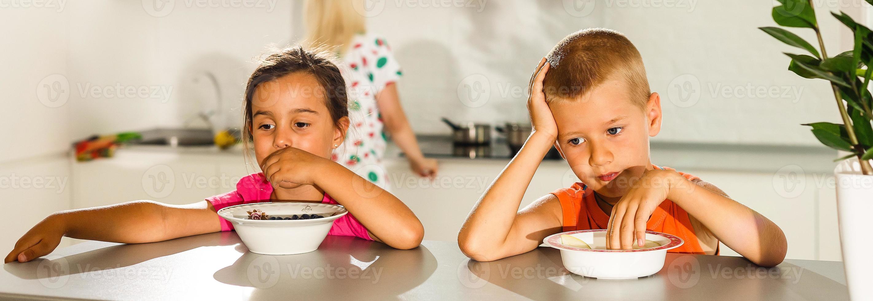 contento joven familia, madre con dos niños, adorable niñito niña y gracioso sucio chico teniendo sano desayuno comiendo Fruta y lácteos, sentado en un blanco soleado cocina con ventana foto