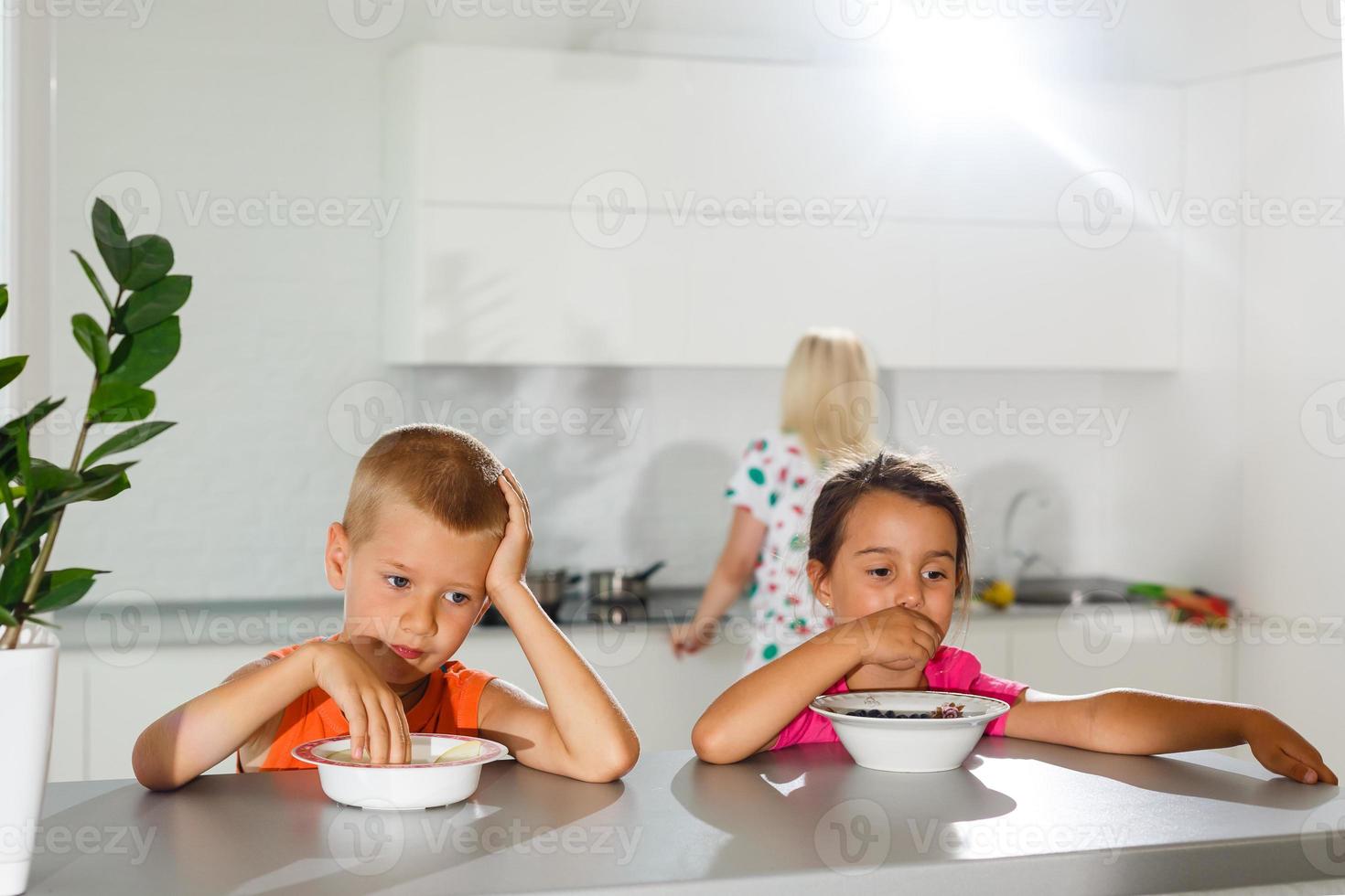 Happy young family, mother with two children, adorable toddler girl and funny messy boy having healthy breakfast eating fruit and dairy, sitting in a white sunny kitchen with window photo