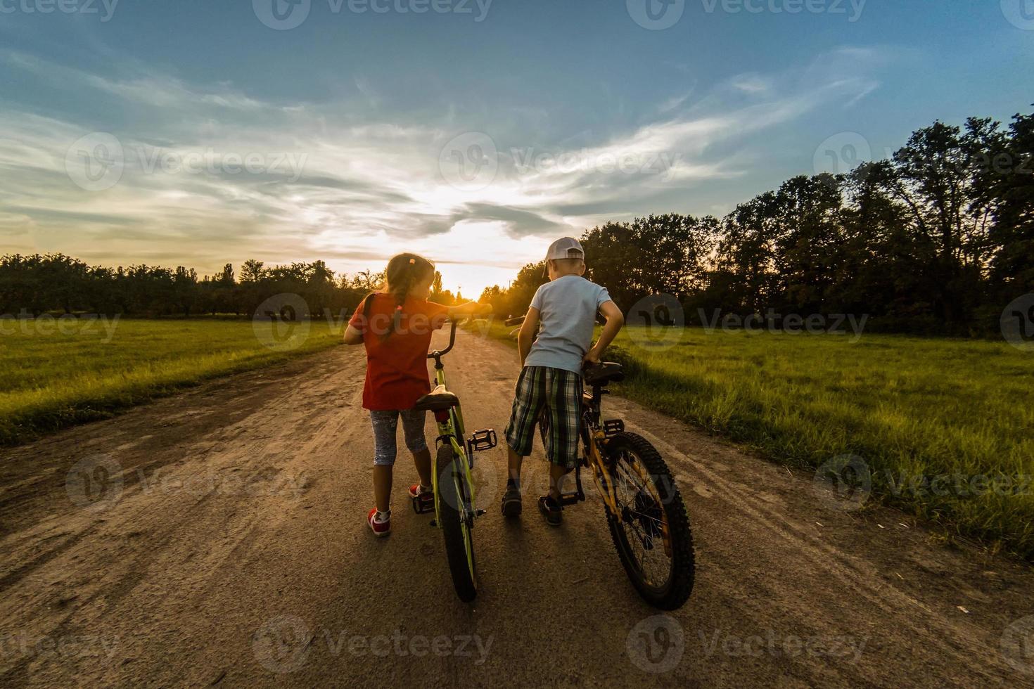 childs riding one bike together on sunny summer evening. sitting on bicycle rack. Family of two people enjoying traveling in scenic field over sunset sky background. photo