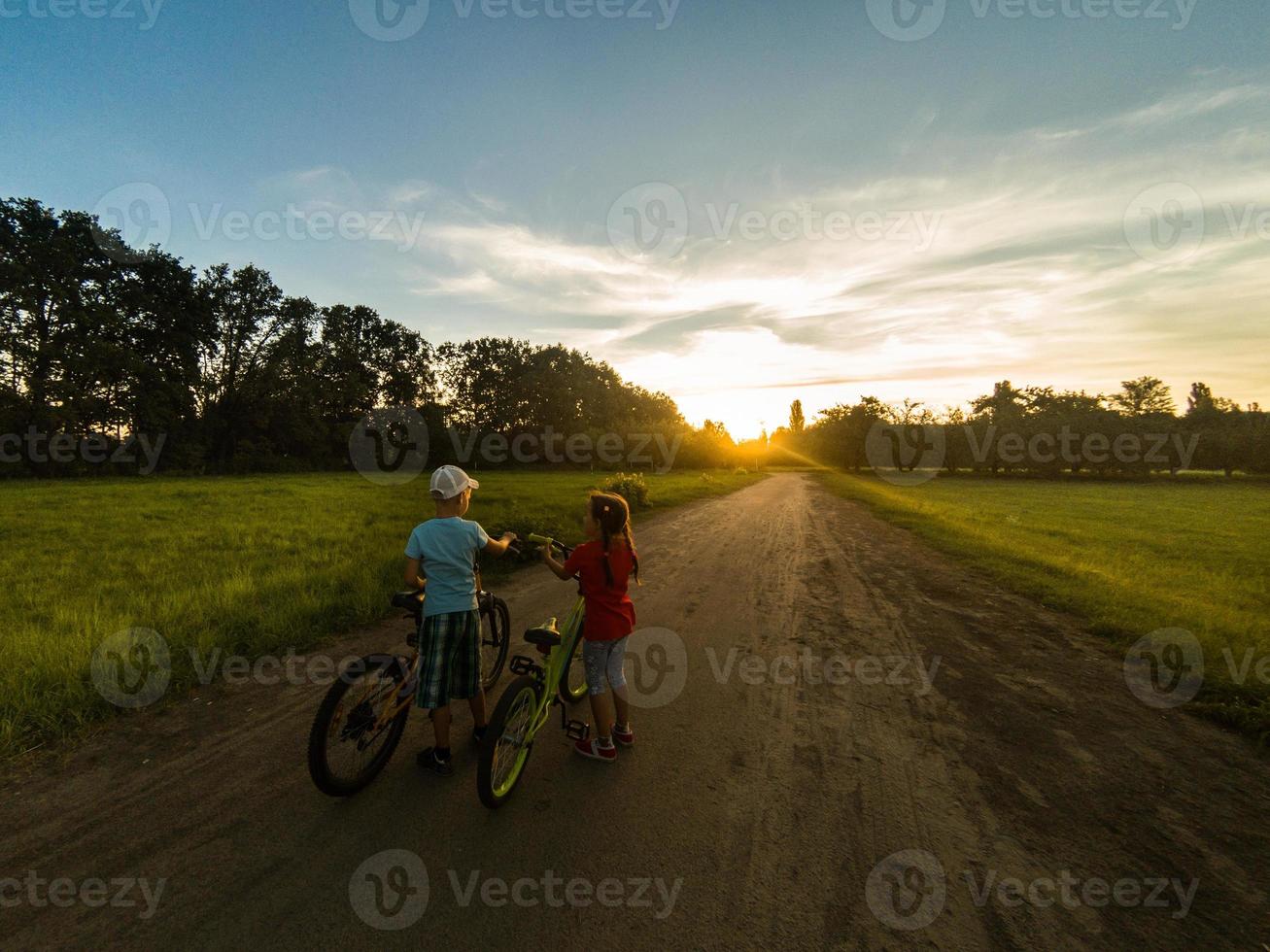 niños montando uno bicicleta juntos en soleado verano noche. sentado en bicicleta estante. familia de dos personas disfrutando de viaje en escénico campo terminado puesta de sol cielo antecedentes. foto