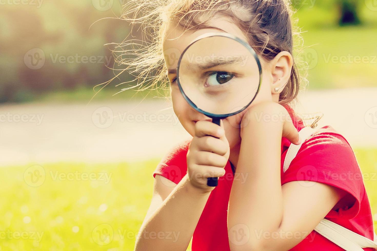 Positive cheerful little girl looking through a magnifying glass outdoors photo