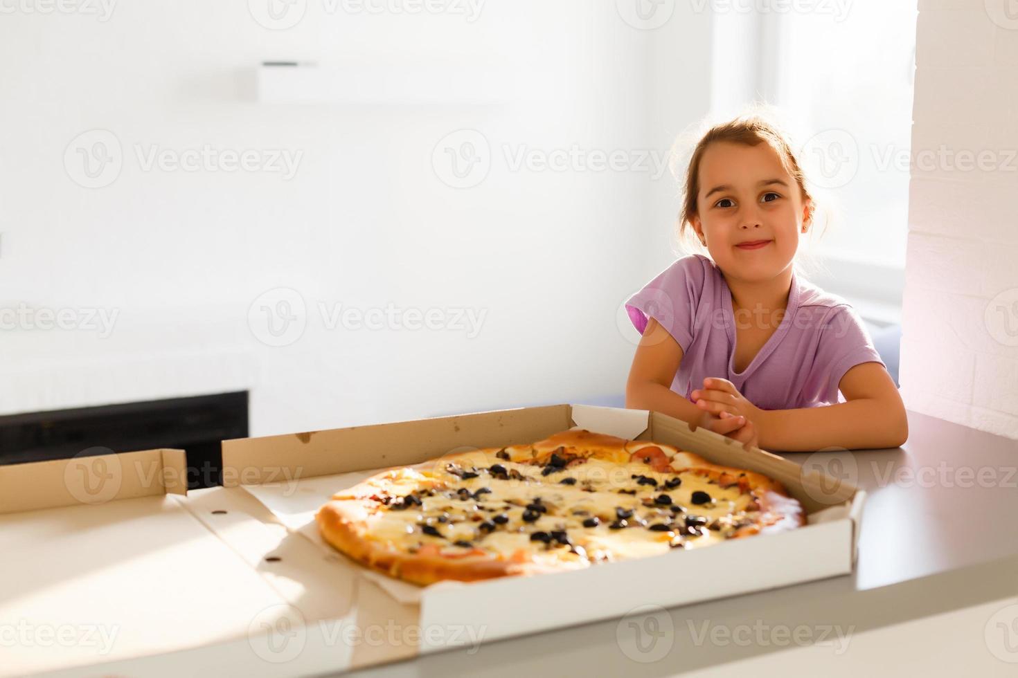 encantador contento joven niña risa y mordiendo apagado grande rebanada de Fresco hecho Pizza. ella sentar a blanco silla en provence estilo interior, sonrisa y disfrutar soleado día y sabroso comida. ella tiene largo rubia cabello. foto