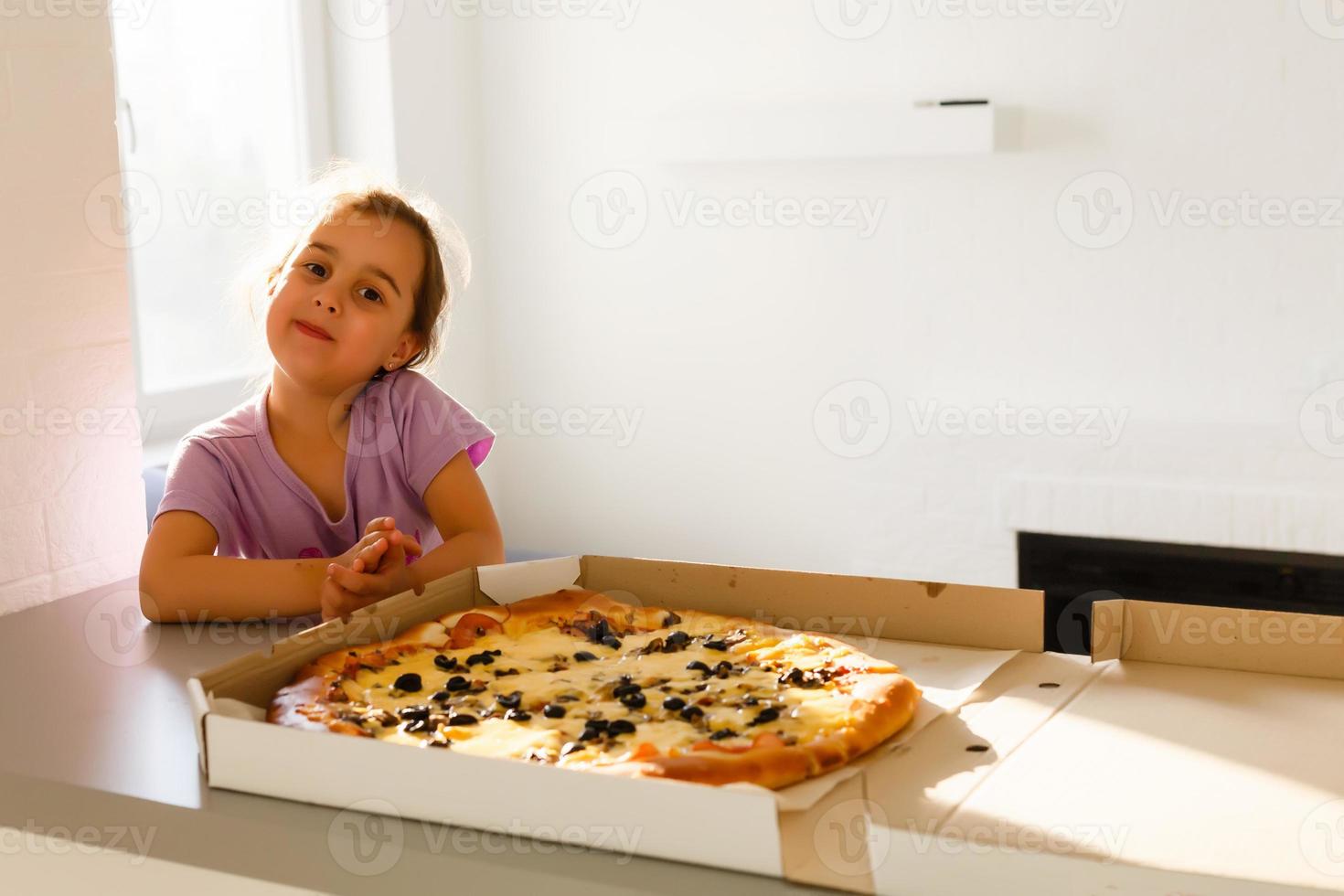 Charming happy young girl laugh and biting off big slice of fresh made pizza. She sit at white chair in Provence style interior, smile and enjoy sunny day and yummy meal. She has long blonde hair. photo