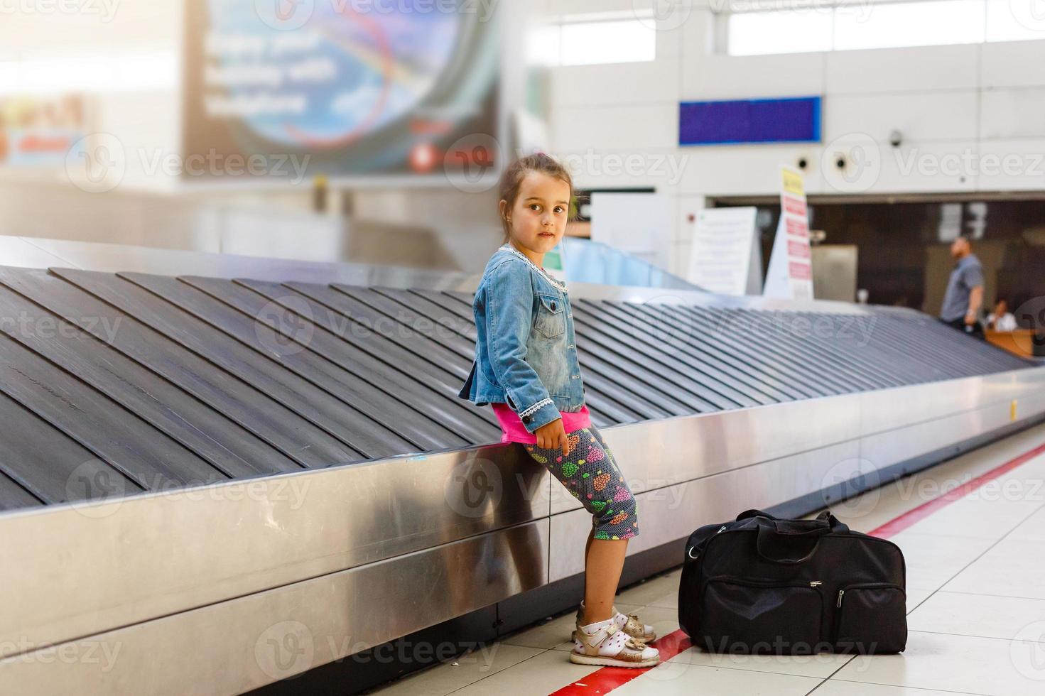 el niña sentado a el aeropuerto en el cinta para equipaje, cuales es situado cerca el recepción. Mira lejos foto
