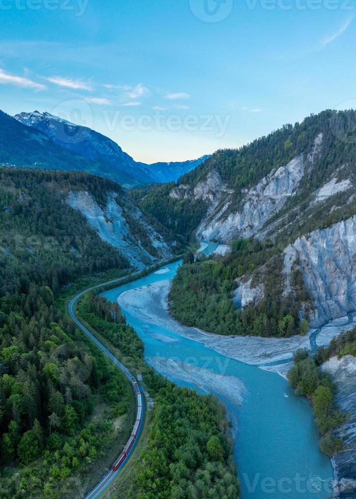 A river that runs in a S curve between the mountains,A train runs next to the river photo