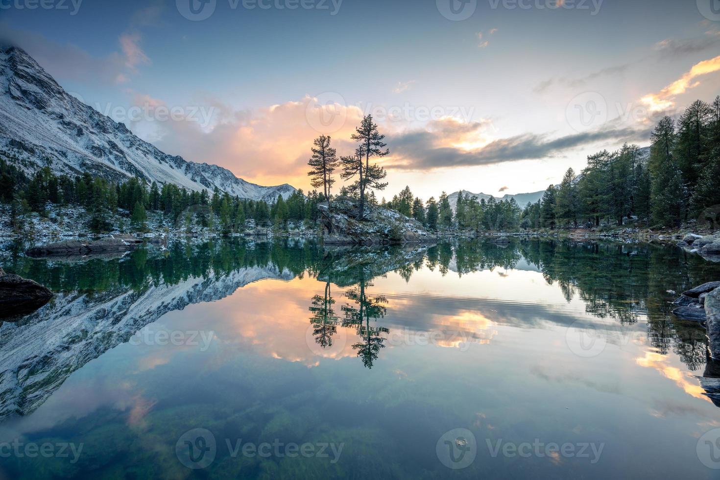 A lake in winter, on which stands a rock with two trees, during a sunset photo