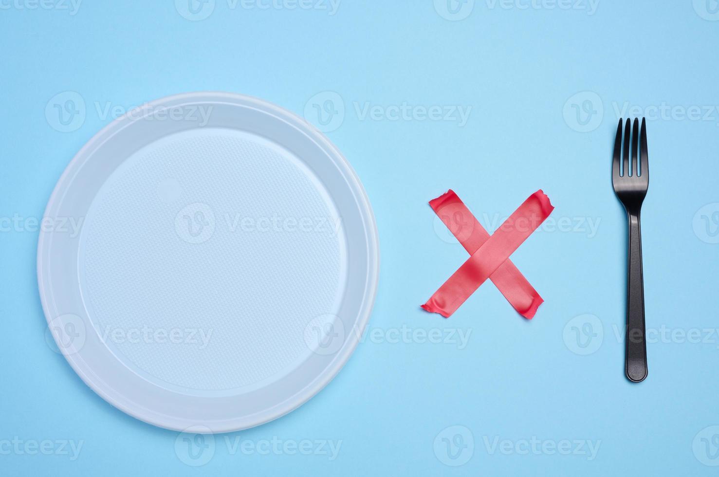 Empty white plastic plate and plastic fork is attached with red adhesive tape on a blue background, a concept of refusing plastic tableware and recycling materials, zero waste photo