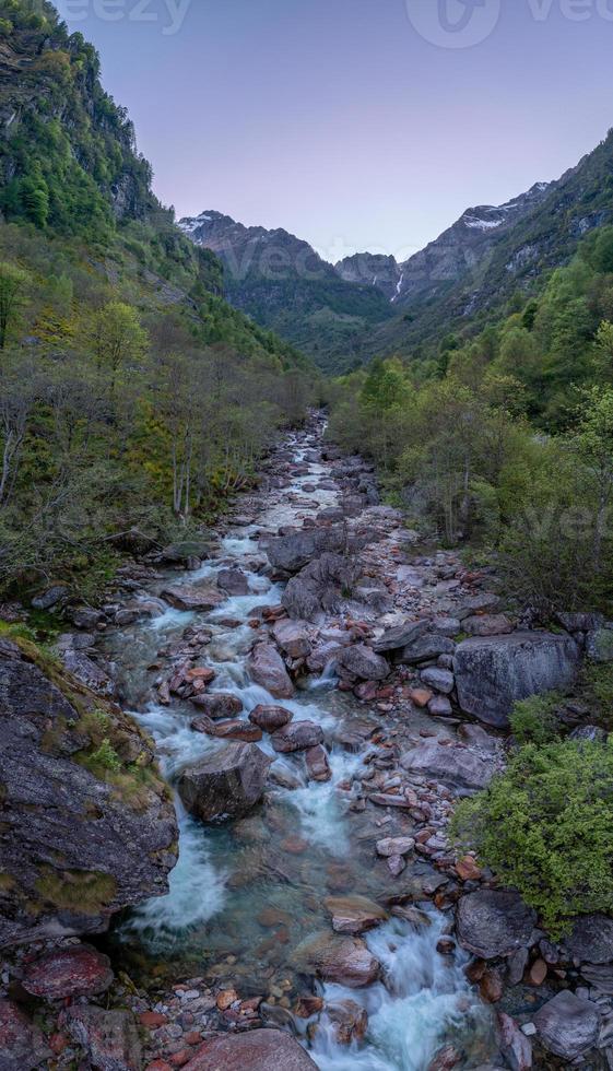 A stream with large boulders that runs between mountains and trees photo