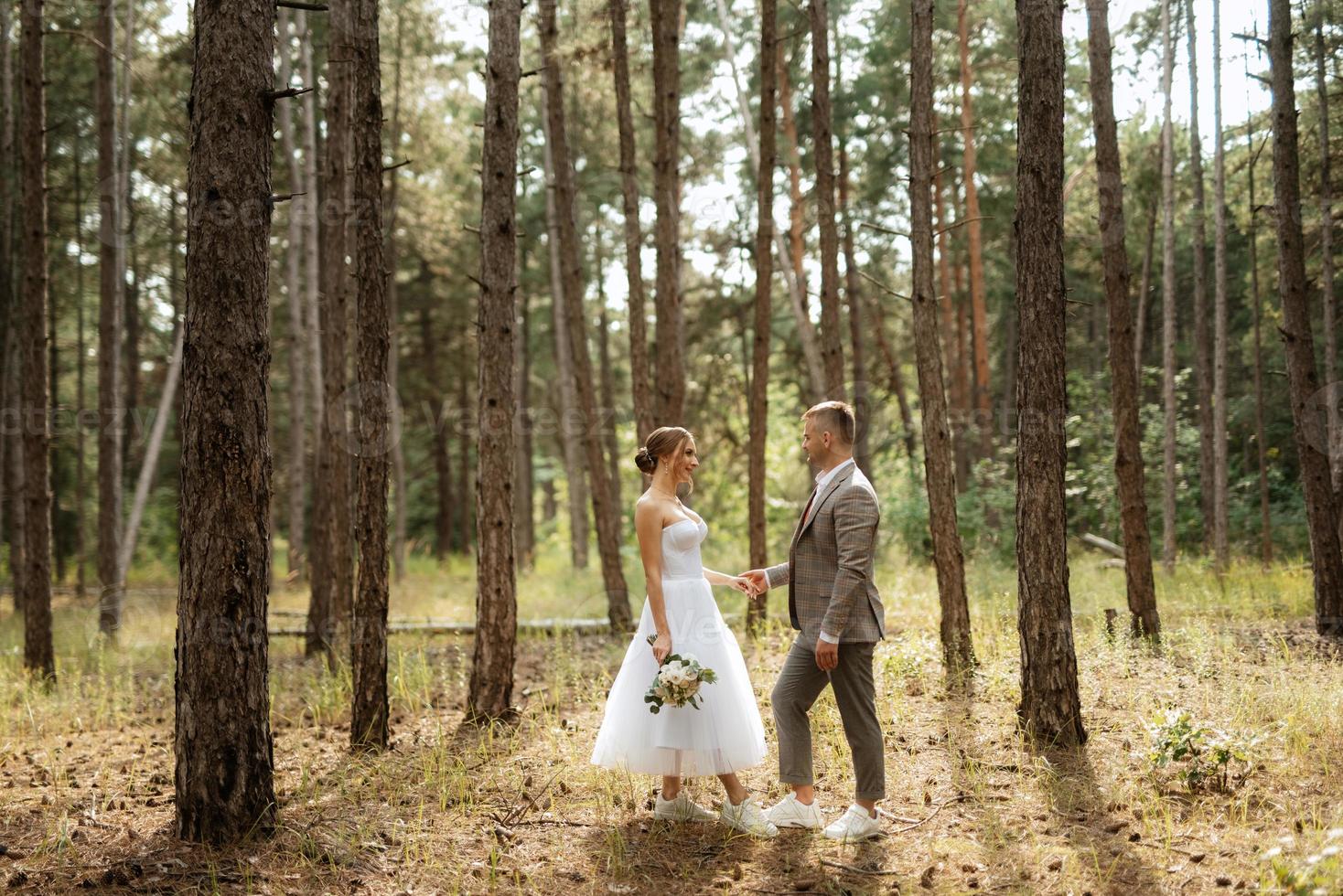 young couple bride in a white short dress and groom in a gray suit in a pine forest photo