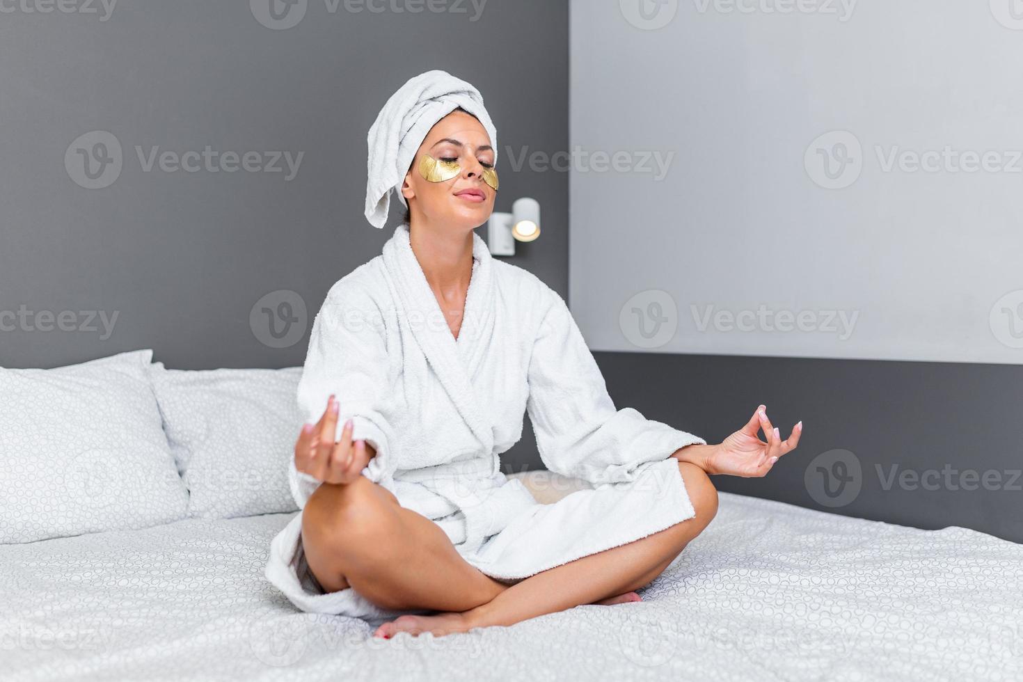 frente ver de pacífico atractivo joven activo mujer practicando yoga en el cama participación ojos cerrado, sentado en medio loto ejercicio, ardha padmasana actitud y vistiendo bata de baño en el Mañana foto