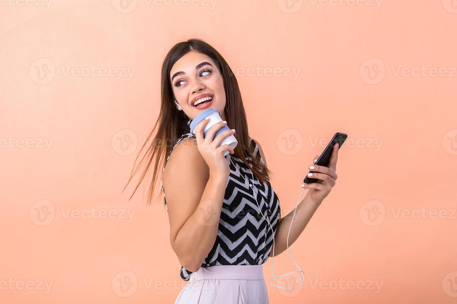 Indoor picture of young good-looking European woman isolated on peach background holding coffe to go and phone in hand and smiling while browsing or reading message photo