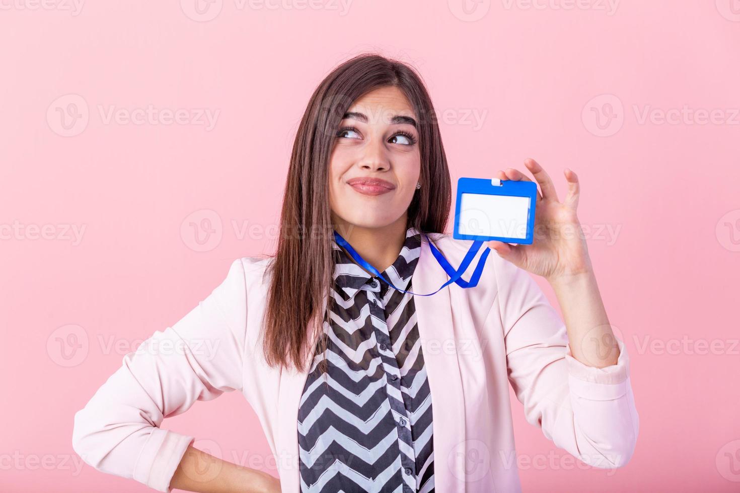 Beautiful success woman shows on camera her badge and smiling over pink wall. Young attractive woman holding blank artist lanyard or badge in hand with metal piece. Plastic pass concept. photo