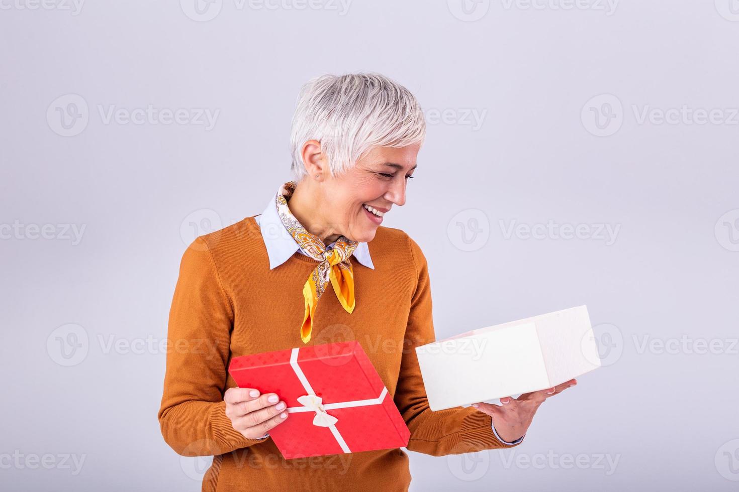 Portrait of a happy smiling mature woman opening a gift box isolated over gray background. Excited senior casual woman holding present box photo