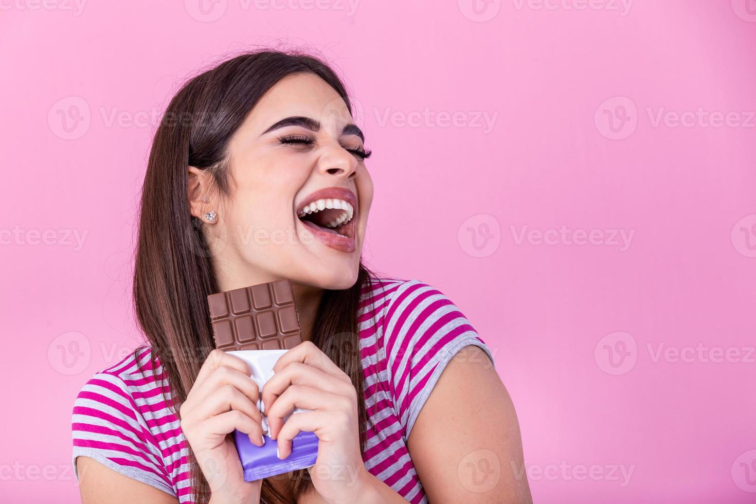 Happy young beautiful lady eating chocolate and smiling. Girl tasting sweet chocolate. Young woman with natural make up having fun and eating chocolate isolated on pink background photo