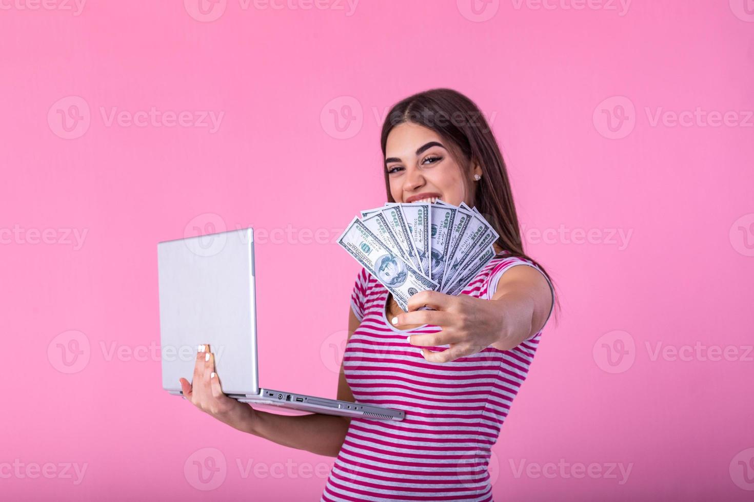 Portrait of an excited satisfied girl holding money banknotes with laptop computer isolated over pink background. Portrait of a cheerful young woman holding money banknotes and laptopcomputer in hands photo