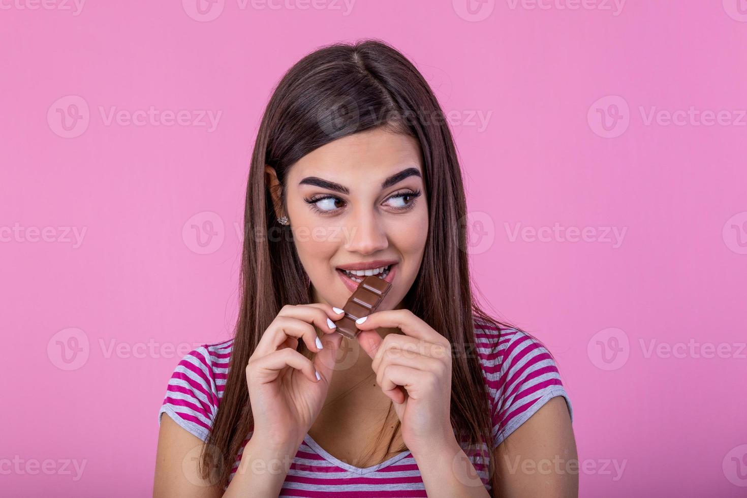 Happy young beautiful lady eating chocolate and smiling. Girl tasting sweet chocolate. Young woman with natural make up having fun and eating chocolate isolated on pink background photo