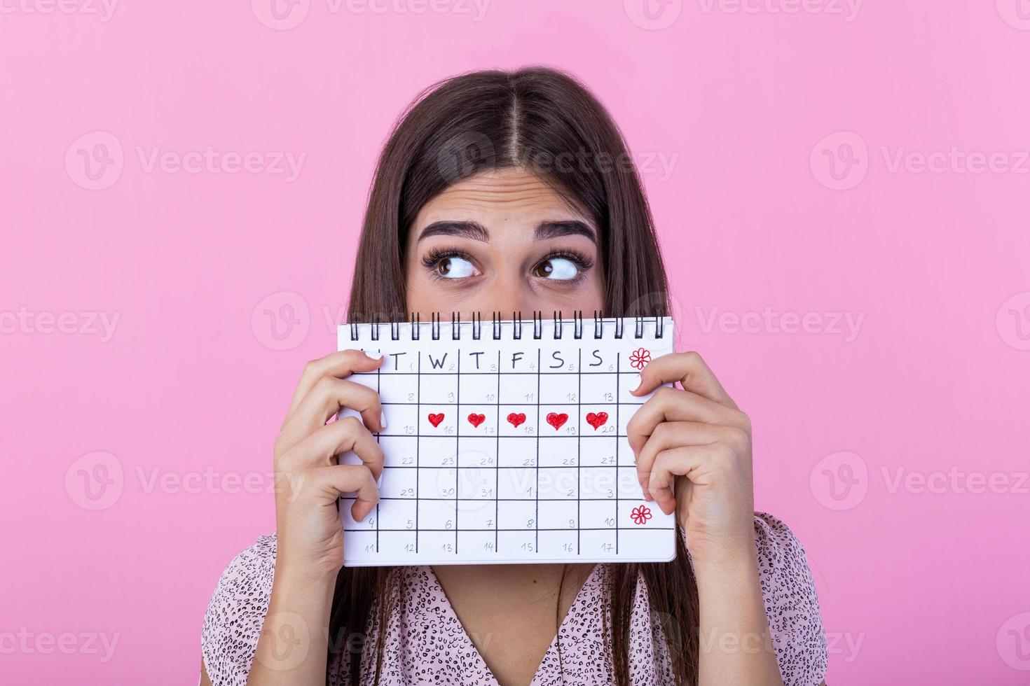 Portrait of a funny young girl in hiding behind a menstrual periods calendar and looking away at copy space isolated over pink background. Female Period calendar photo
