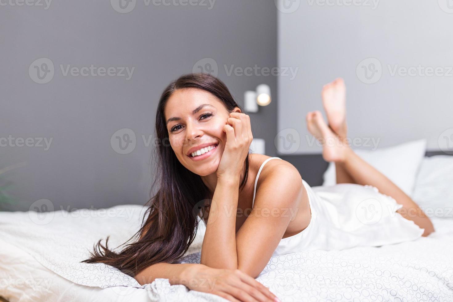 alegre mujer acostado en el cama a hogar . hermosa sonriente mujer con Fresco suave piel y natural maquillaje en ropa interior teniendo divertido acostado en blanco cama. sano contento hembra modelo relajante adentro. foto