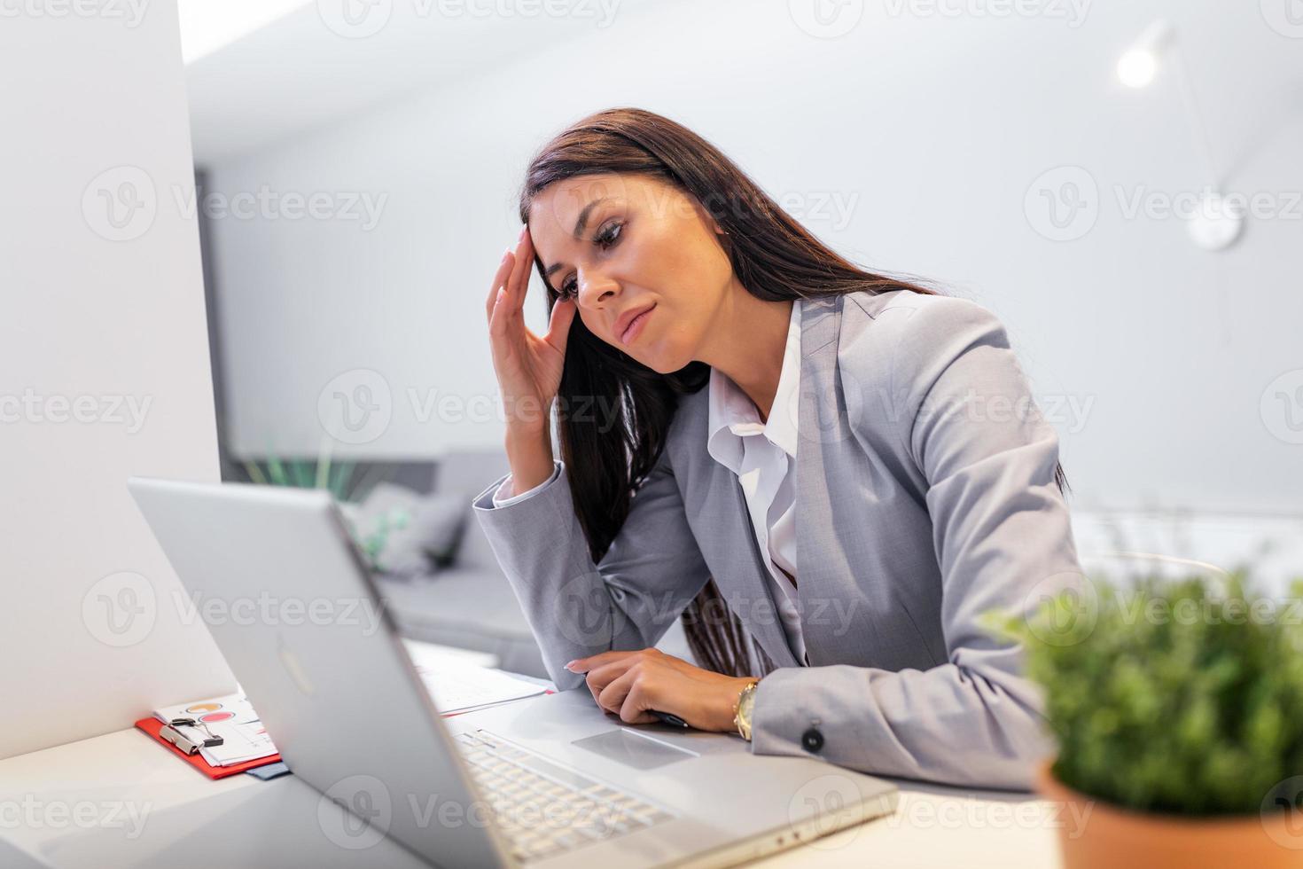 Young frustrated woman working at office desk in front of laptop suffering from chronic daily headaches, treatment online, appointing to a medical consultation, electromagnetic radiation, sick pay photo