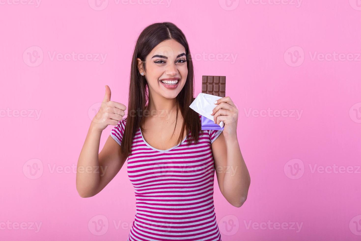 Portrait of a happy young woman with chocolate bar isolated over pink background showing thumbs up. Young woman with natural make up having fun and eating chocolate isolated on pink background photo