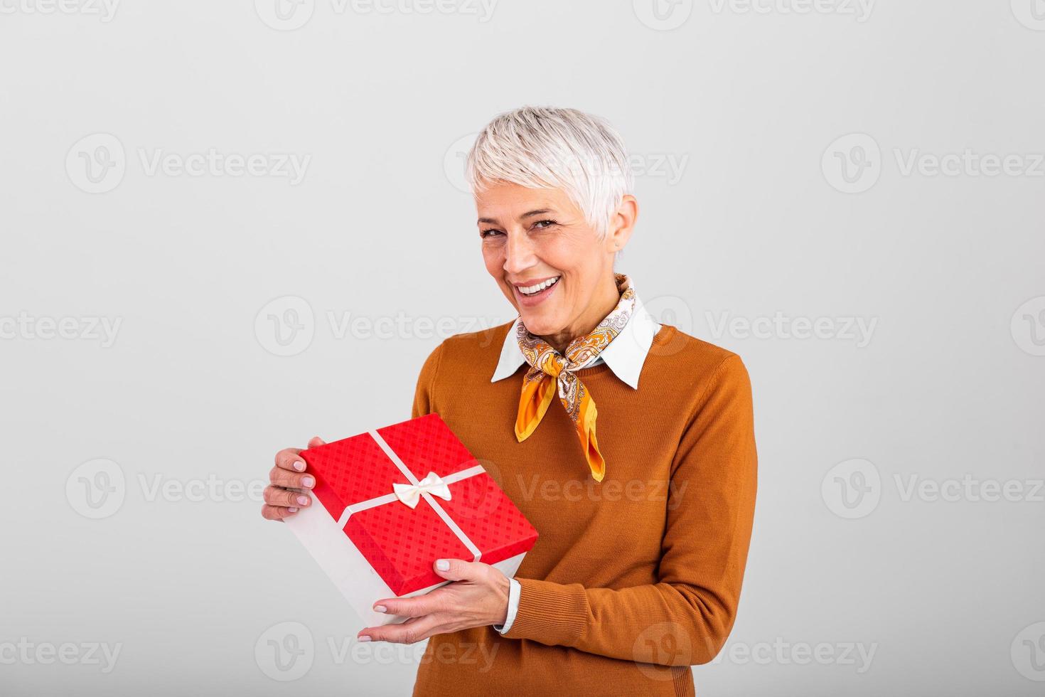 Cheerful good looking senior woman holding a red Christmas gift box in her hands, against a gray background. Concept holidays and presents photo
