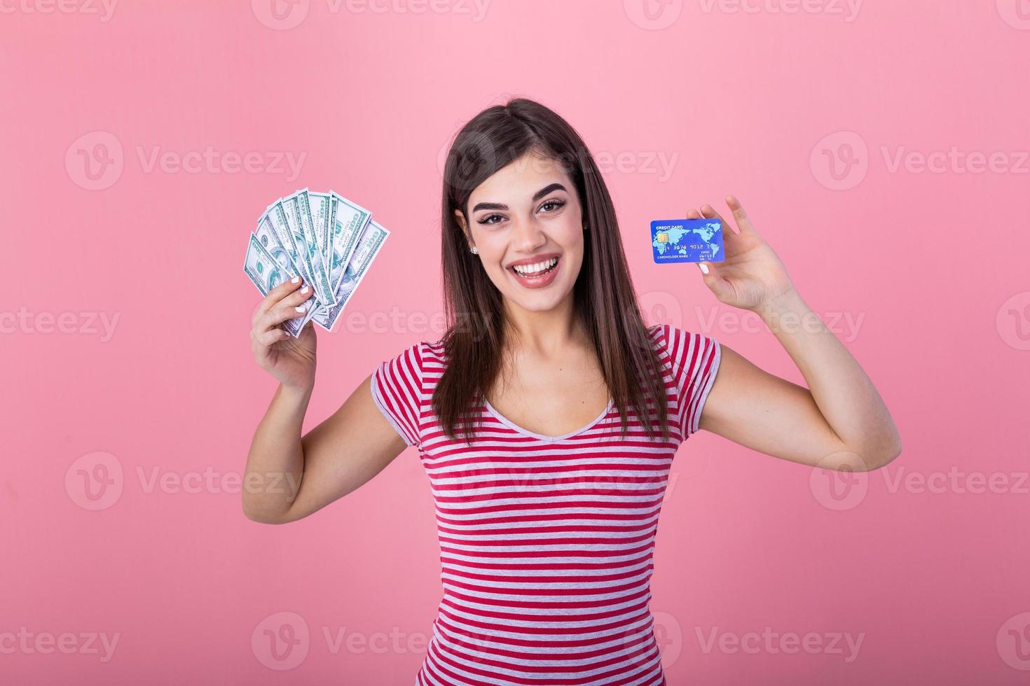 Portrait of a girl in a stripe t-shirt standing on a pink background. Model looks at the camera showing bank card and a bundle of dollars with dreamy look photo