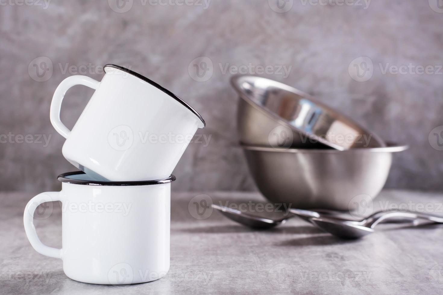 Two white empty metal mugs and metal bowls and spoons on a gray background photo