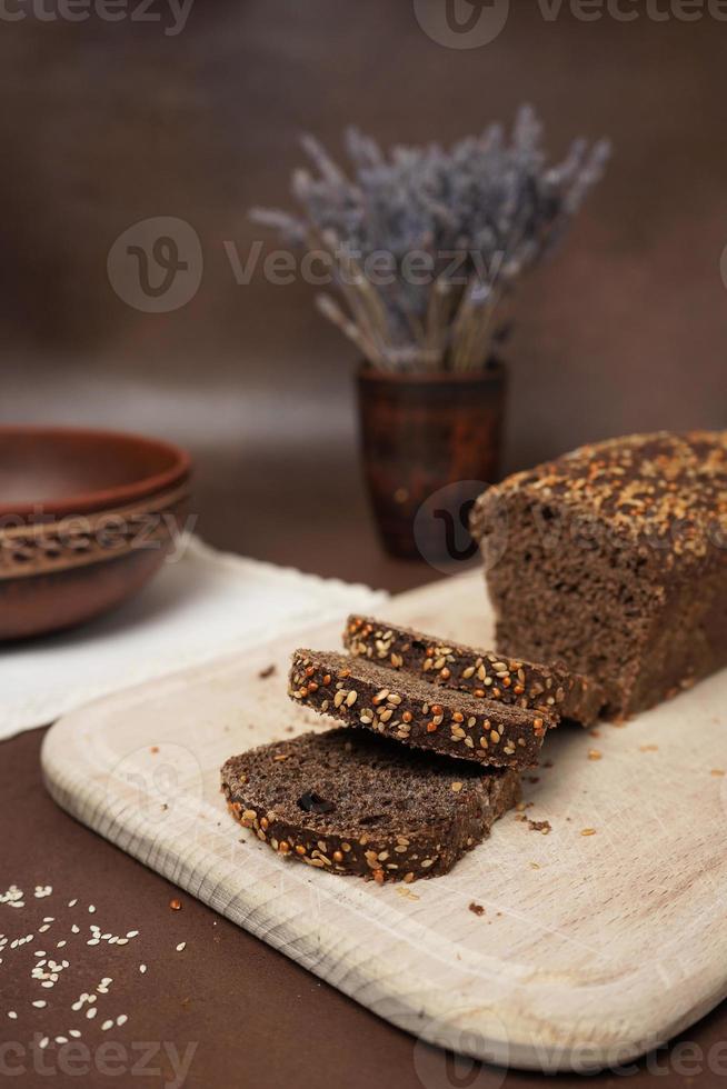 Black Bread with cereals Sliced on a Wooden cutting Close up Board against a brown background. Next to the bread is a Clay Plate, fork, spoon and white cotton tablecloth photo