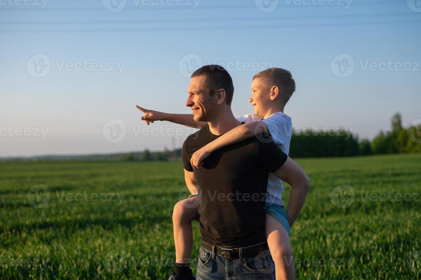 The boy is sitting on his dad's back pointing his finger to the side and they laugh photo