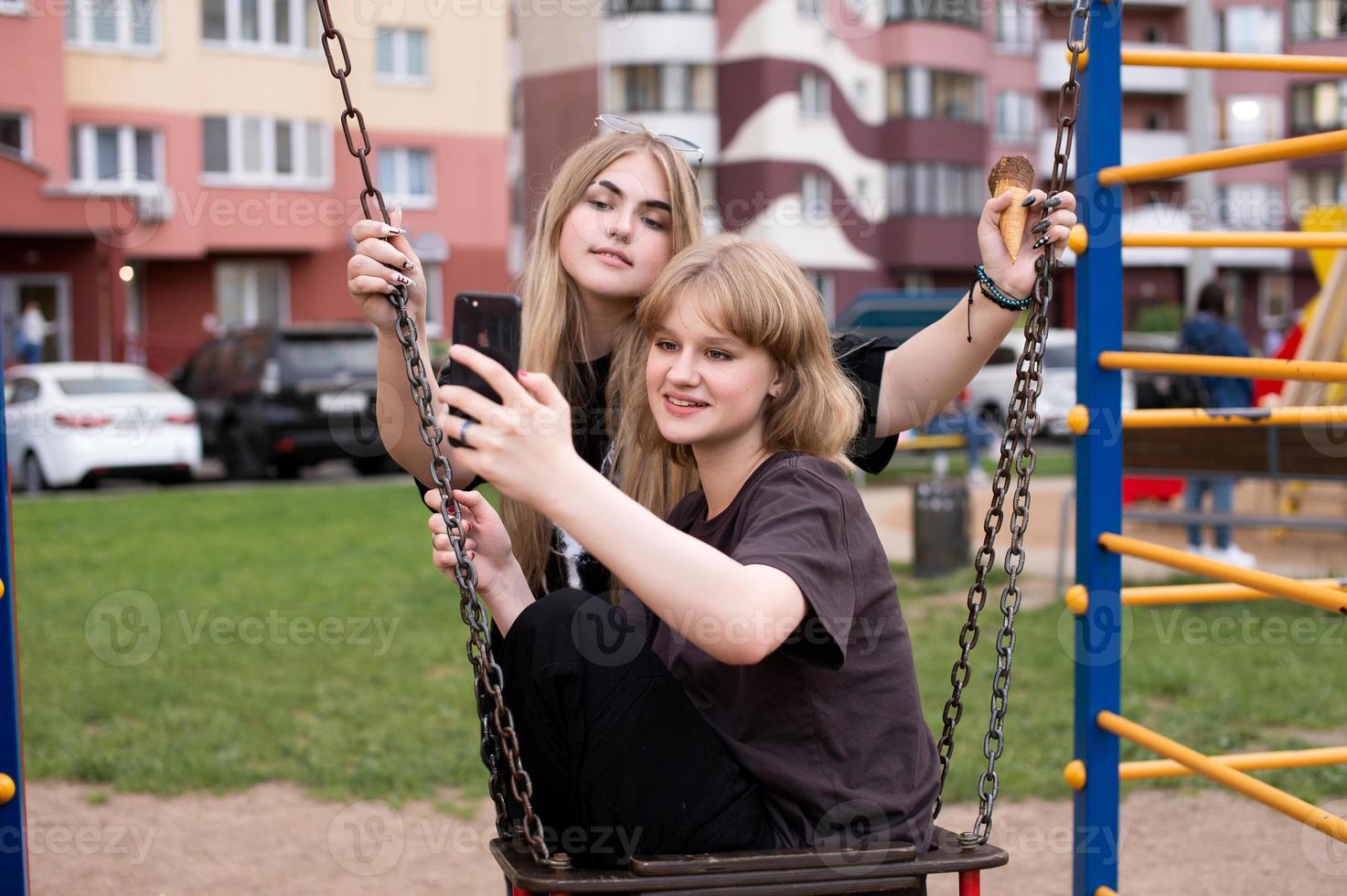 Two funny girls are sitting on a swing in the city and taking selfies on their phone. Teenagers smile photo