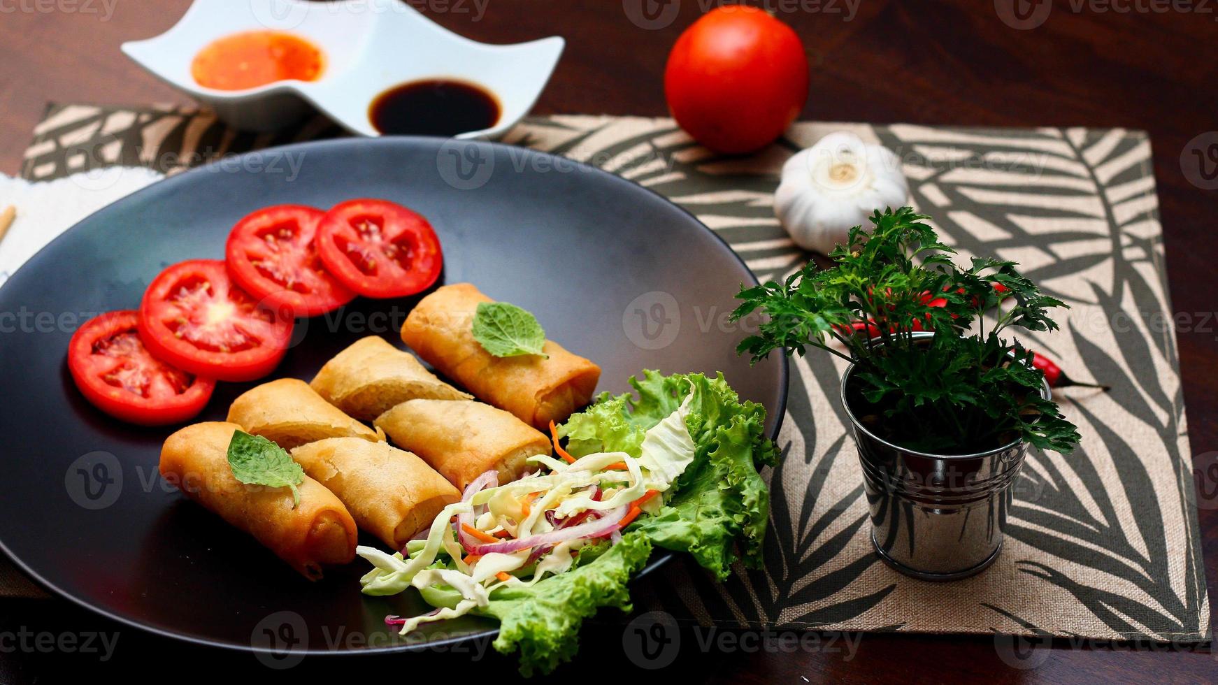Fried spring rolls with vegetables and tomatoes placed in a black plate on a black wooden table and dipping sauce. photo