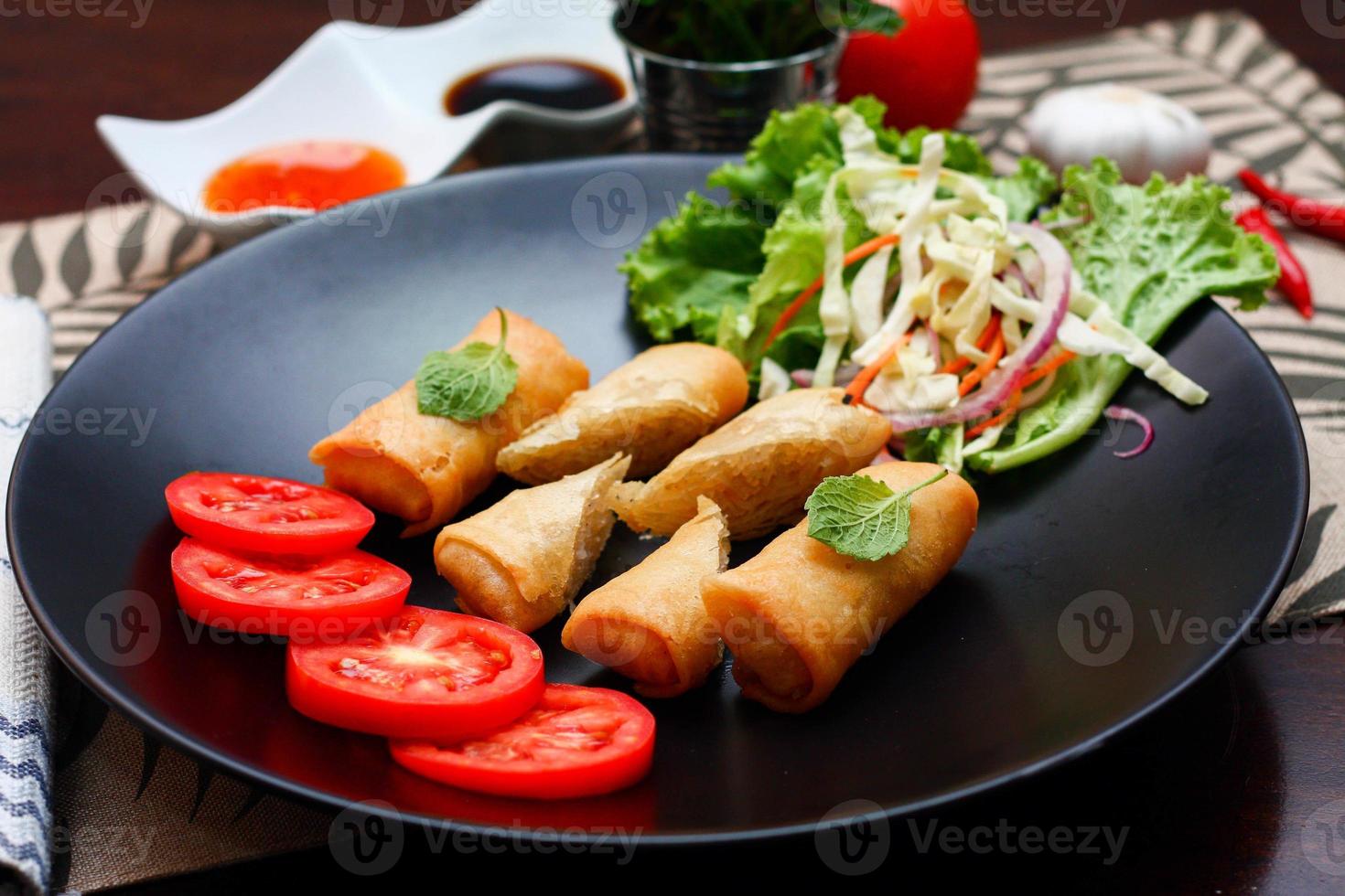 Fried spring rolls with vegetables and tomatoes placed in a black plate on a black wooden table and dipping sauce. photo