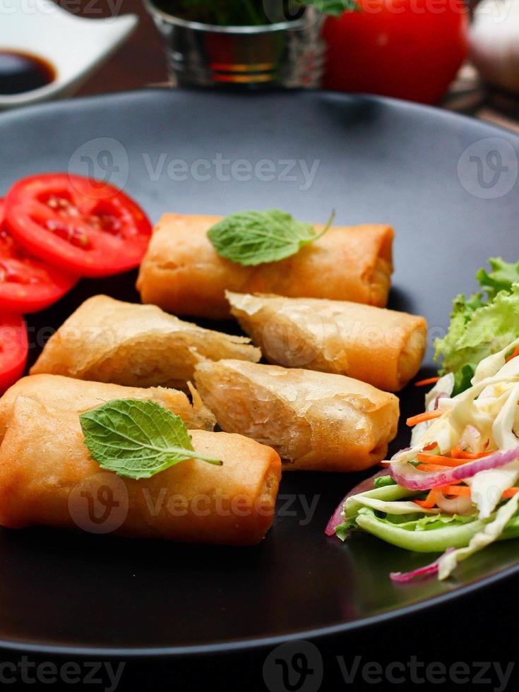 frito primavera rollos con vegetales y Tomates metido en un negro plato en un negro de madera mesa y inmersión salsa. foto