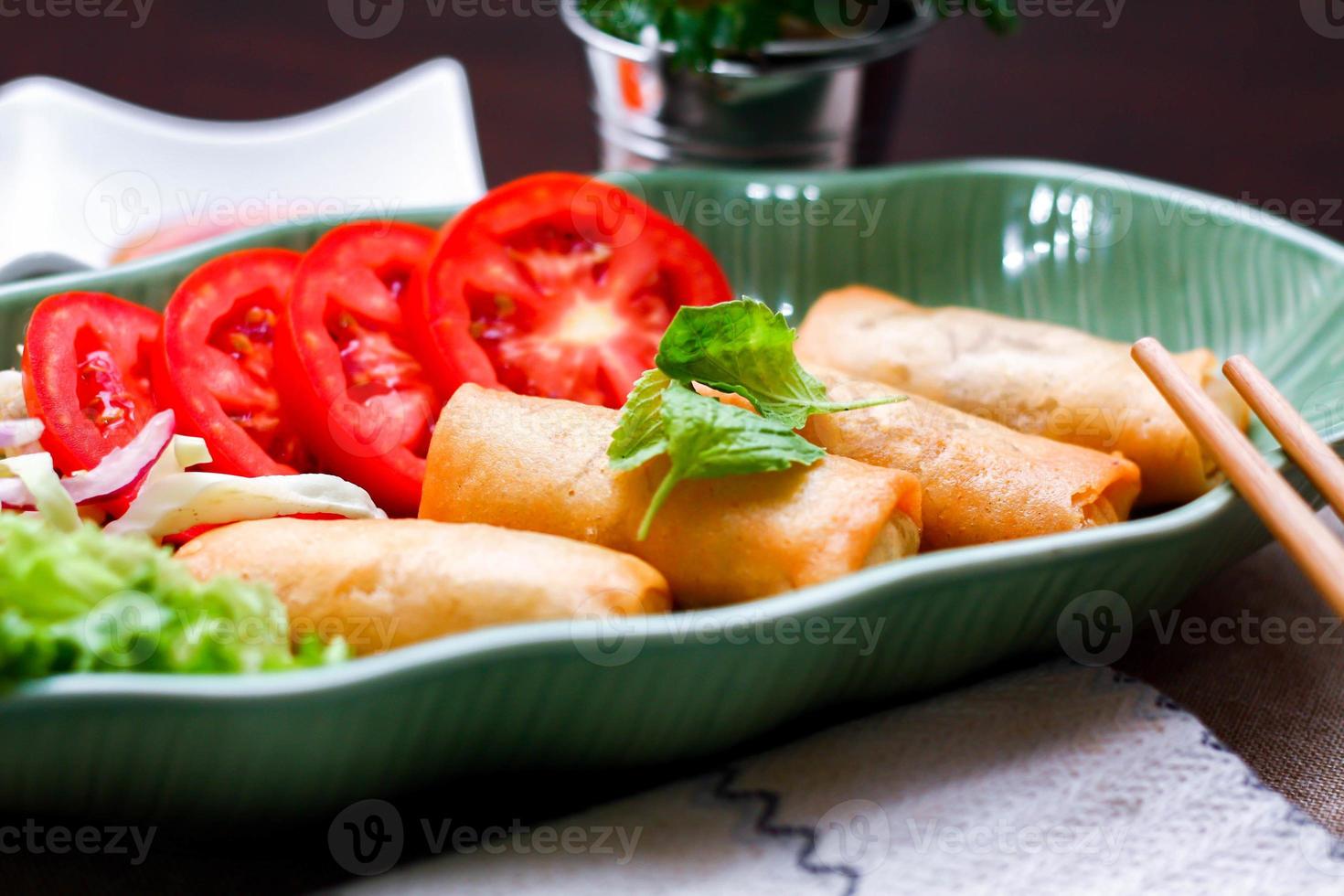 Fried spring rolls, vegetables and tomatoes placed in a green leaf shape plate on a black wooden table and dipping sauce. photo