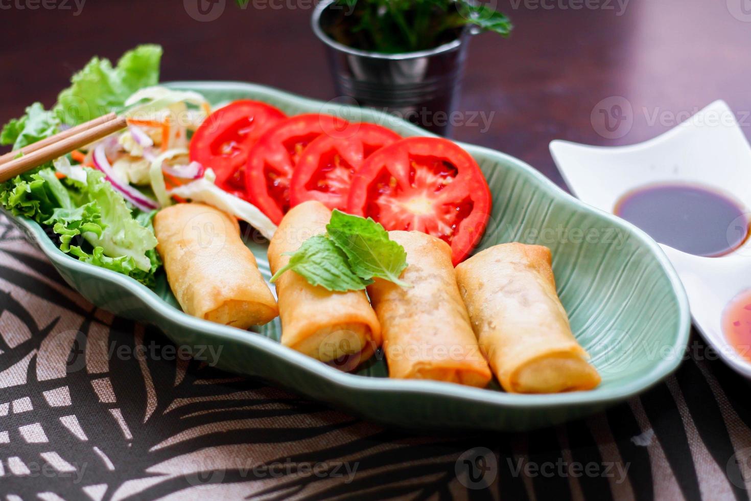 Fried spring rolls, vegetables and tomatoes placed in a green leaf shape plate on a black wooden table and dipping sauce. photo