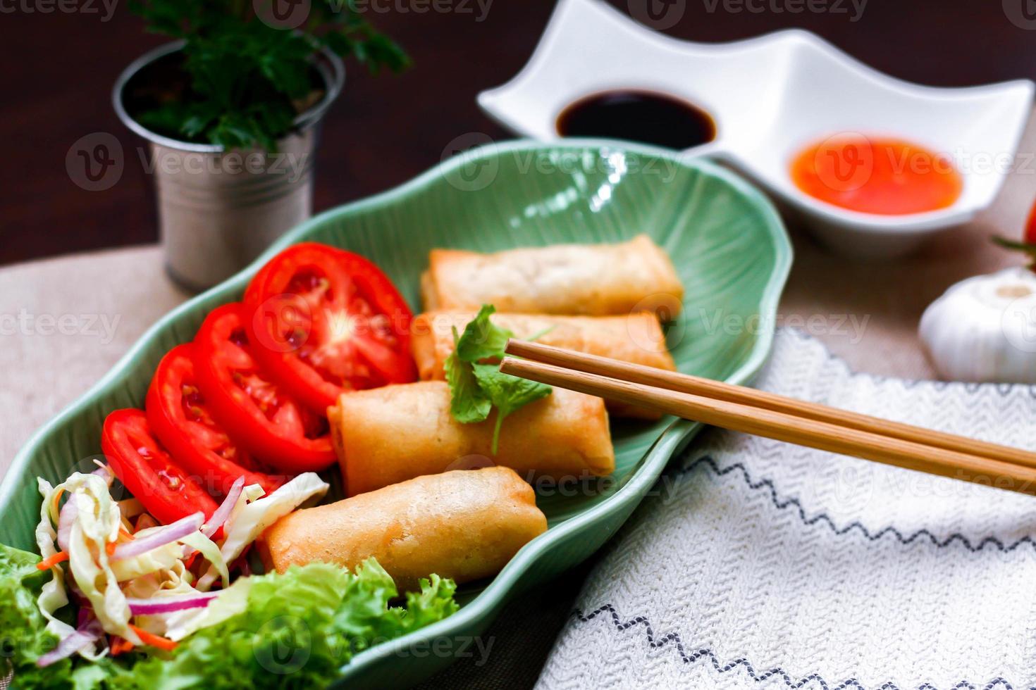Fried spring rolls, vegetables and tomatoes placed in a green leaf shape plate on a black wooden table and dipping sauce. photo