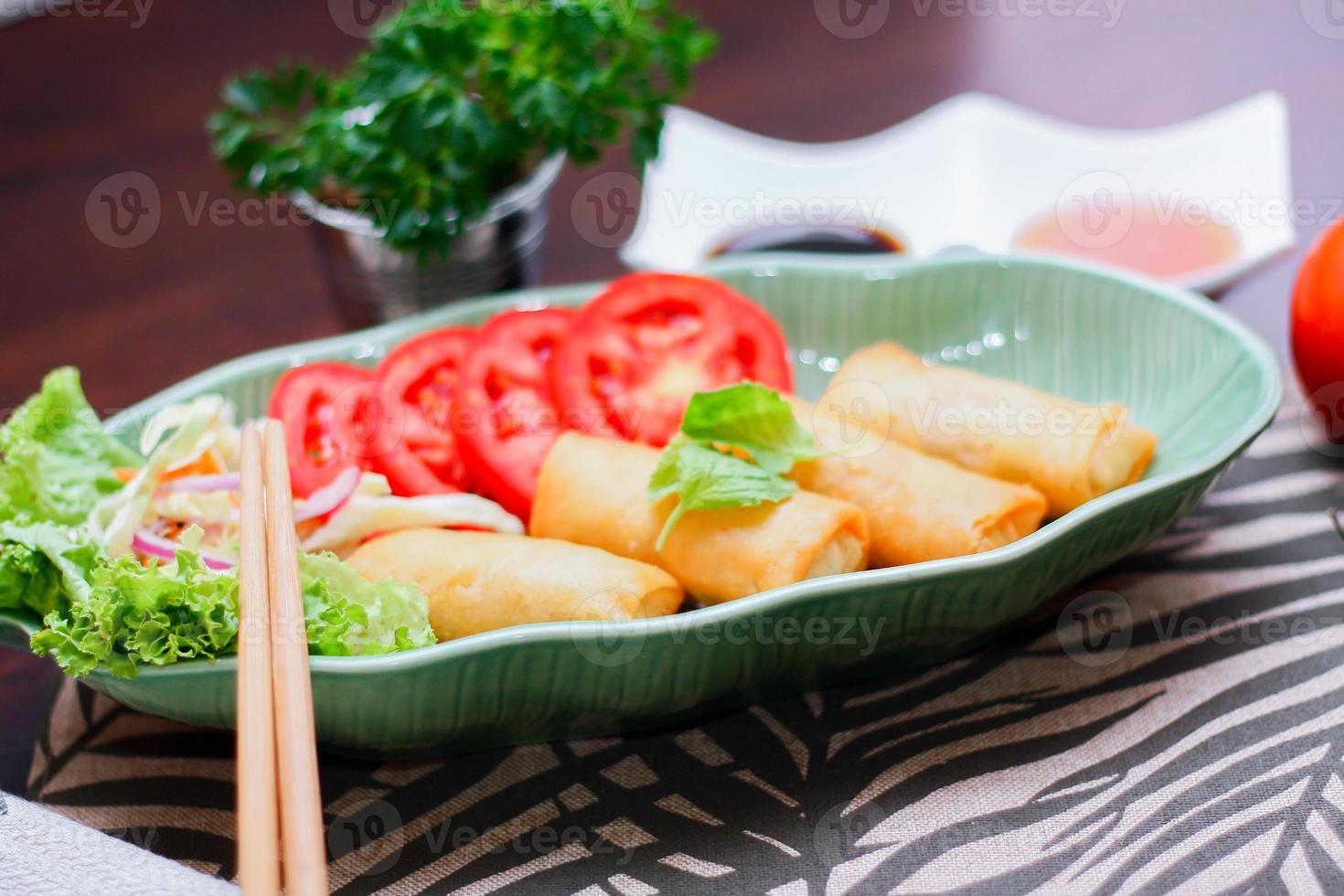 Fried spring rolls, vegetables and tomatoes placed in a green leaf shape plate on a black wooden table and dipping sauce. photo