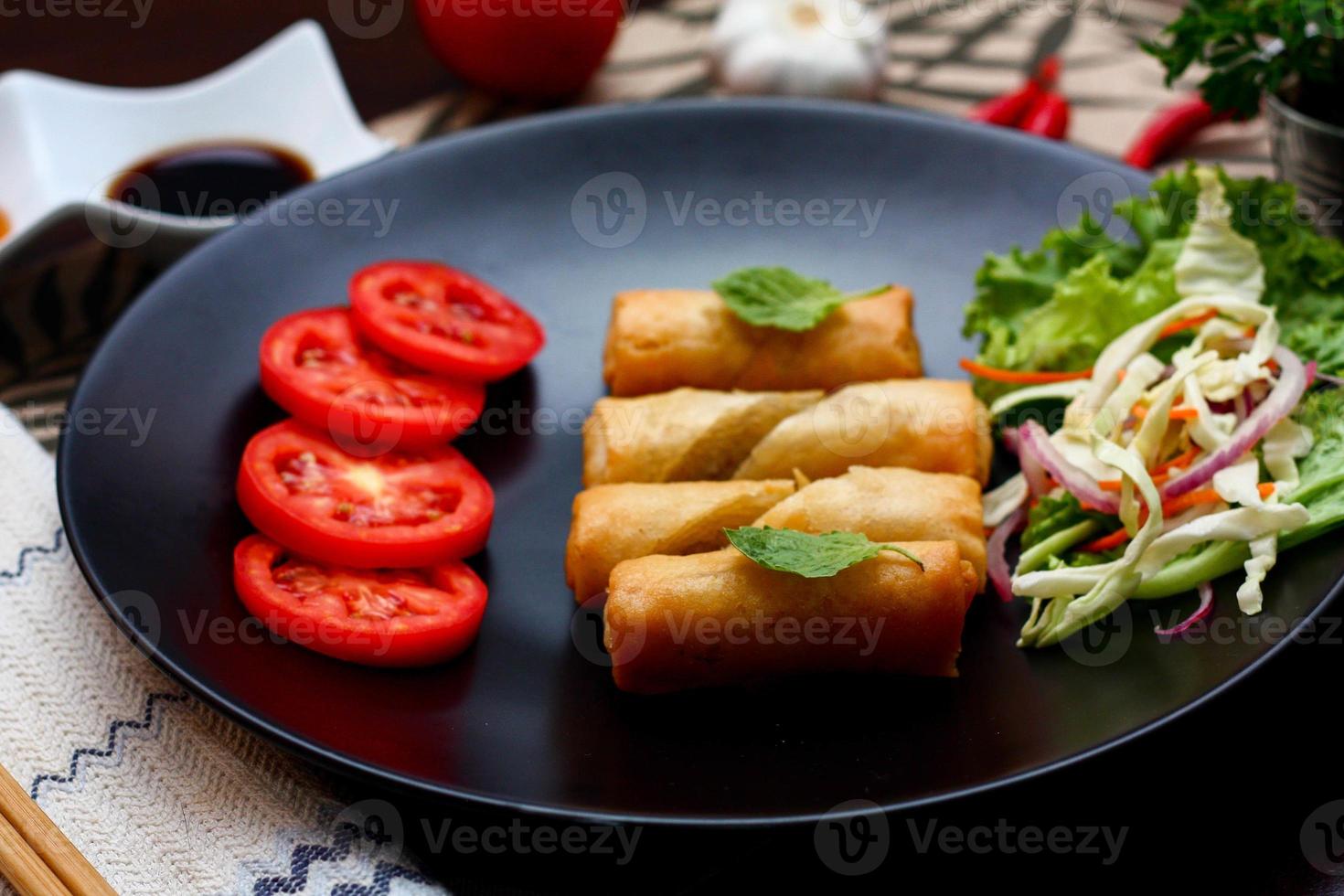 Fried spring rolls with vegetables and tomatoes placed in a black plate on a black wooden table and dipping sauce. photo