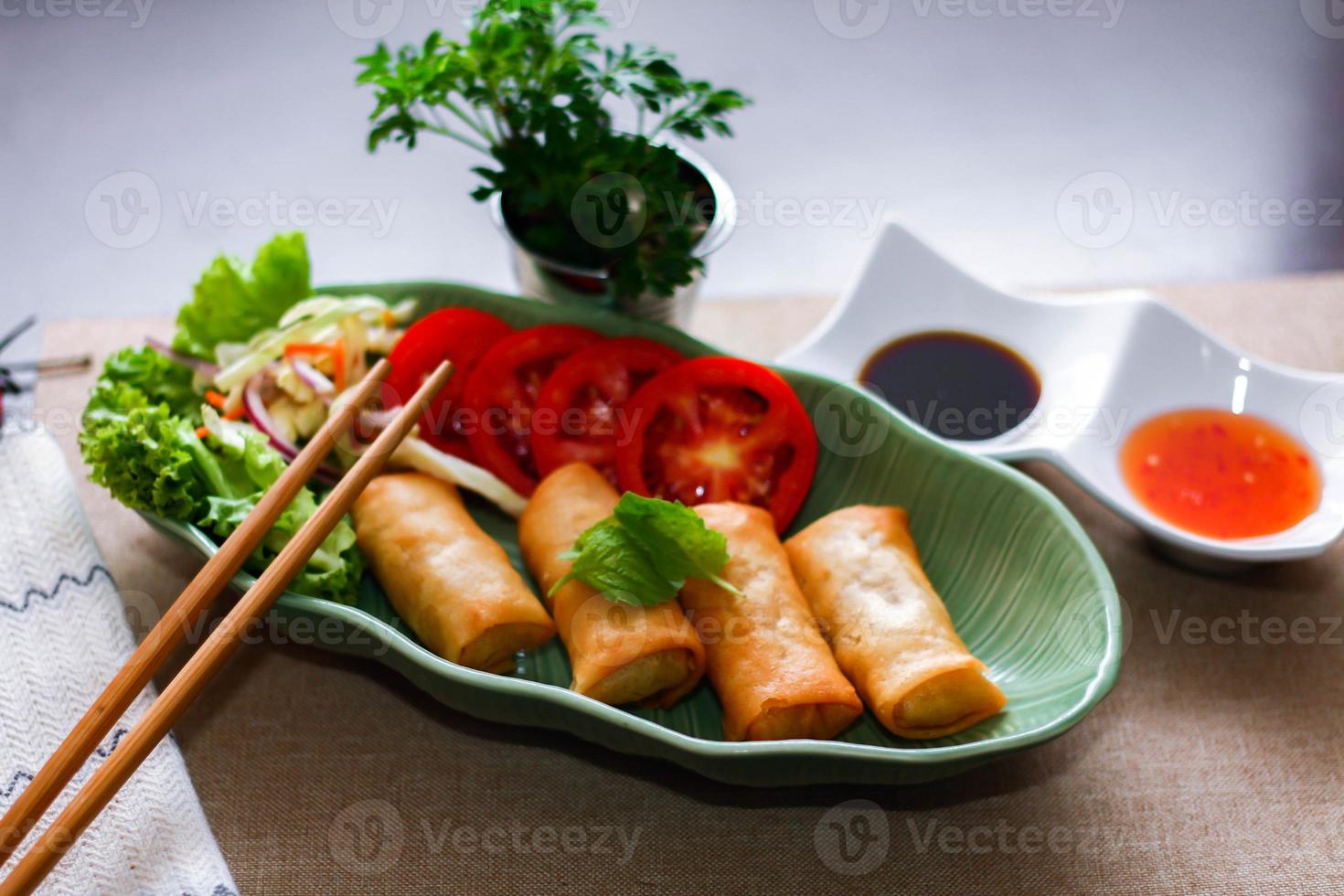 Fried spring rolls, vegetables and tomatoes placed in a green leaf shape plate on a black wooden table and dipping sauce. photo