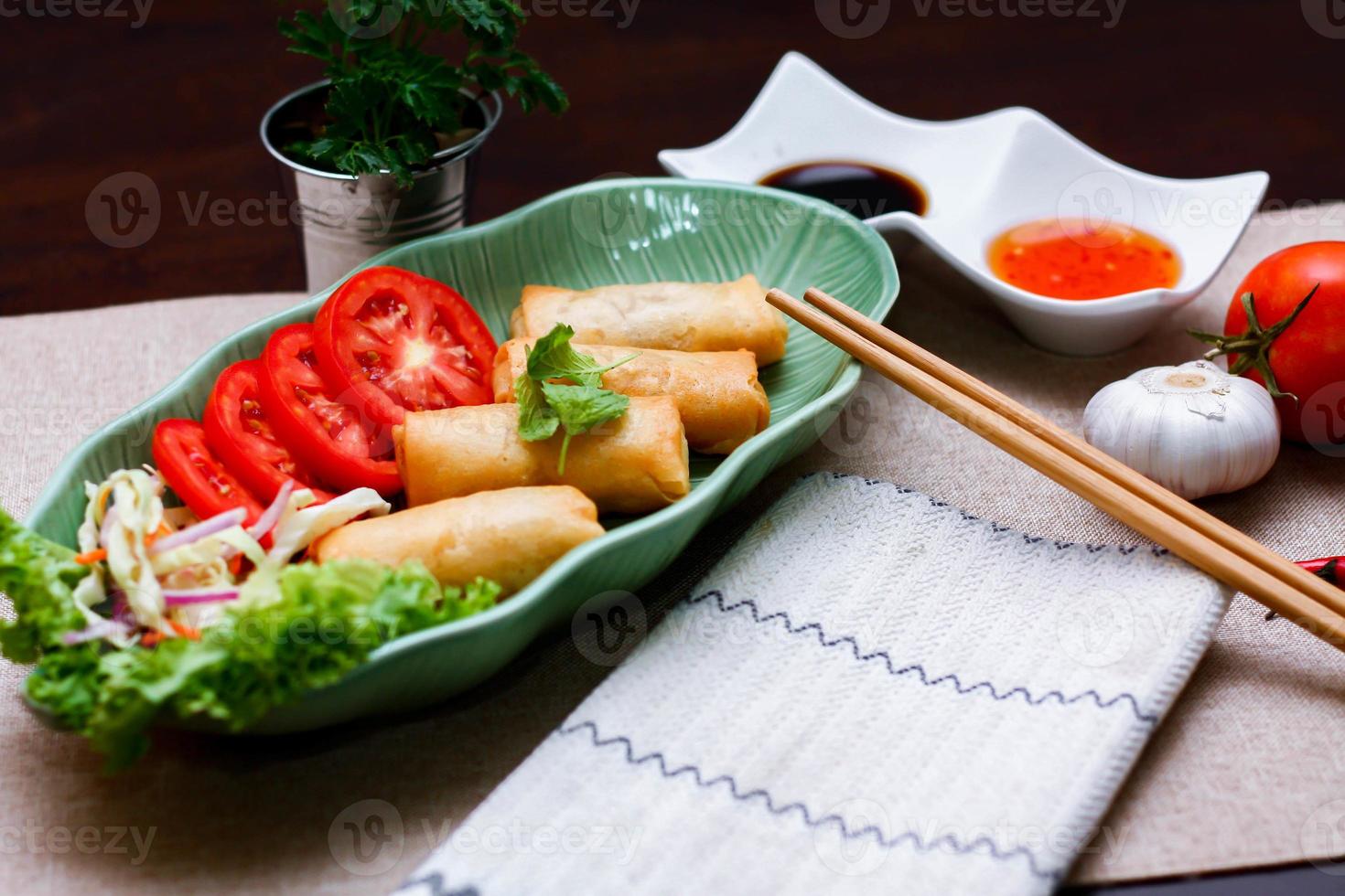 Fried spring rolls, vegetables and tomatoes placed in a green leaf shape plate on a black wooden table and dipping sauce. photo