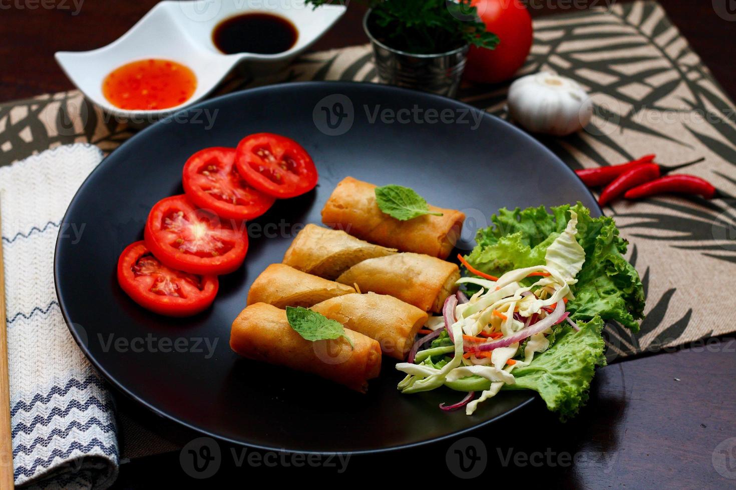 frito primavera rollos con vegetales y Tomates metido en un negro plato en un negro de madera mesa y inmersión salsa. foto