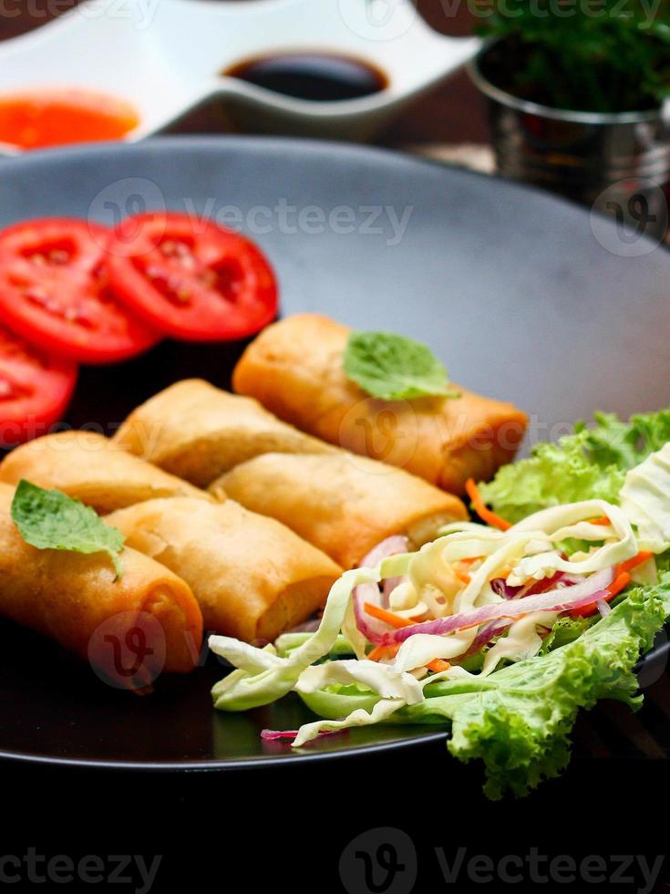 Fried spring rolls with vegetables and tomatoes placed in a black plate on a black wooden table and dipping sauce. photo