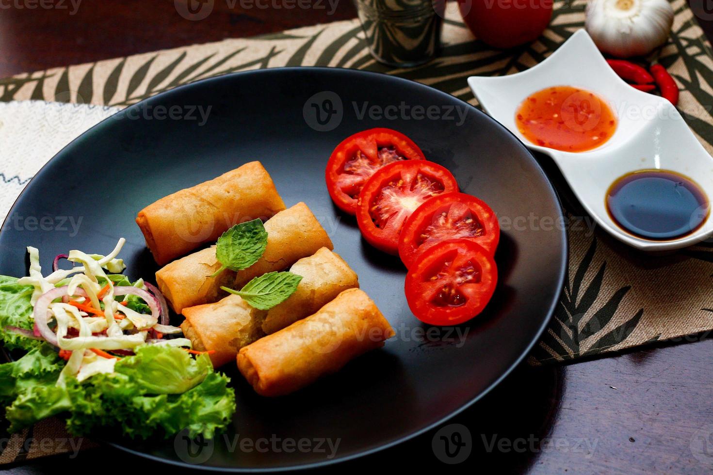 Fried spring rolls with vegetables and tomatoes placed in a black plate on a black wooden table and dipping sauce. photo