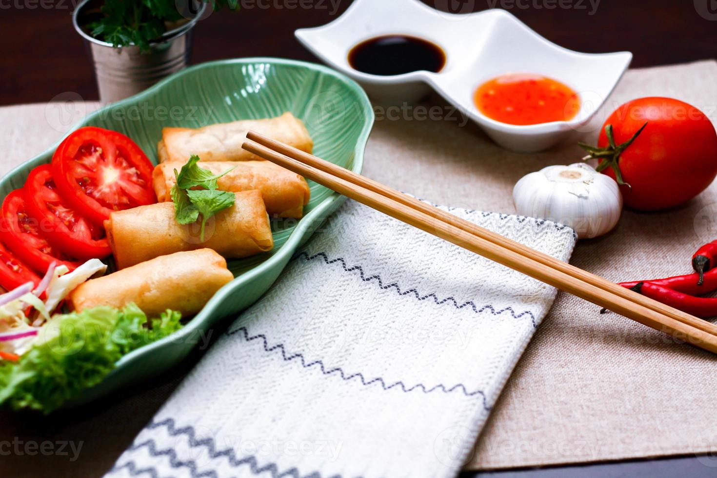 Fried spring rolls, vegetables and tomatoes placed in a green leaf shape plate on a black wooden table and dipping sauce. photo