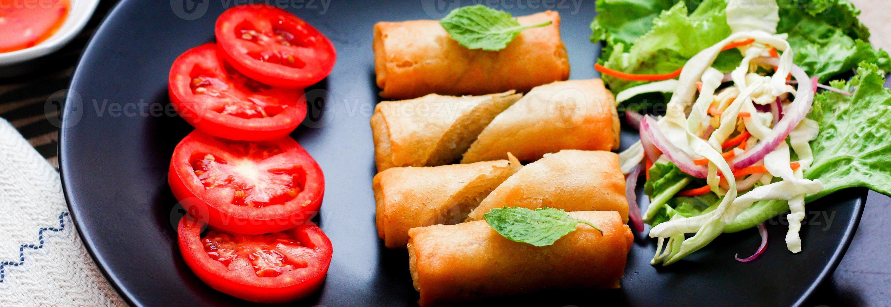 frito primavera rollos con vegetales y Tomates metido en un negro plato en un negro de madera mesa y inmersión salsa. foto