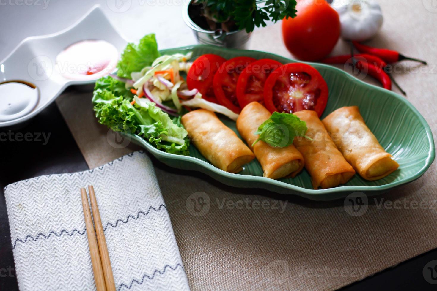 Fried spring rolls, vegetables and tomatoes placed in a green leaf shape plate on a black wooden table and dipping sauce. photo