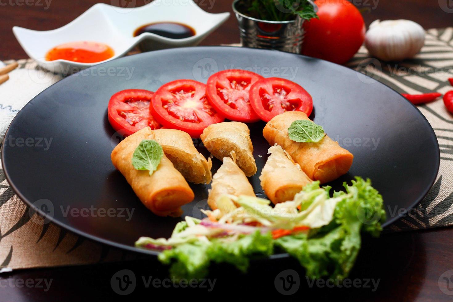 Fried spring rolls with vegetables and tomatoes placed in a black plate on a black wooden table and dipping sauce. photo